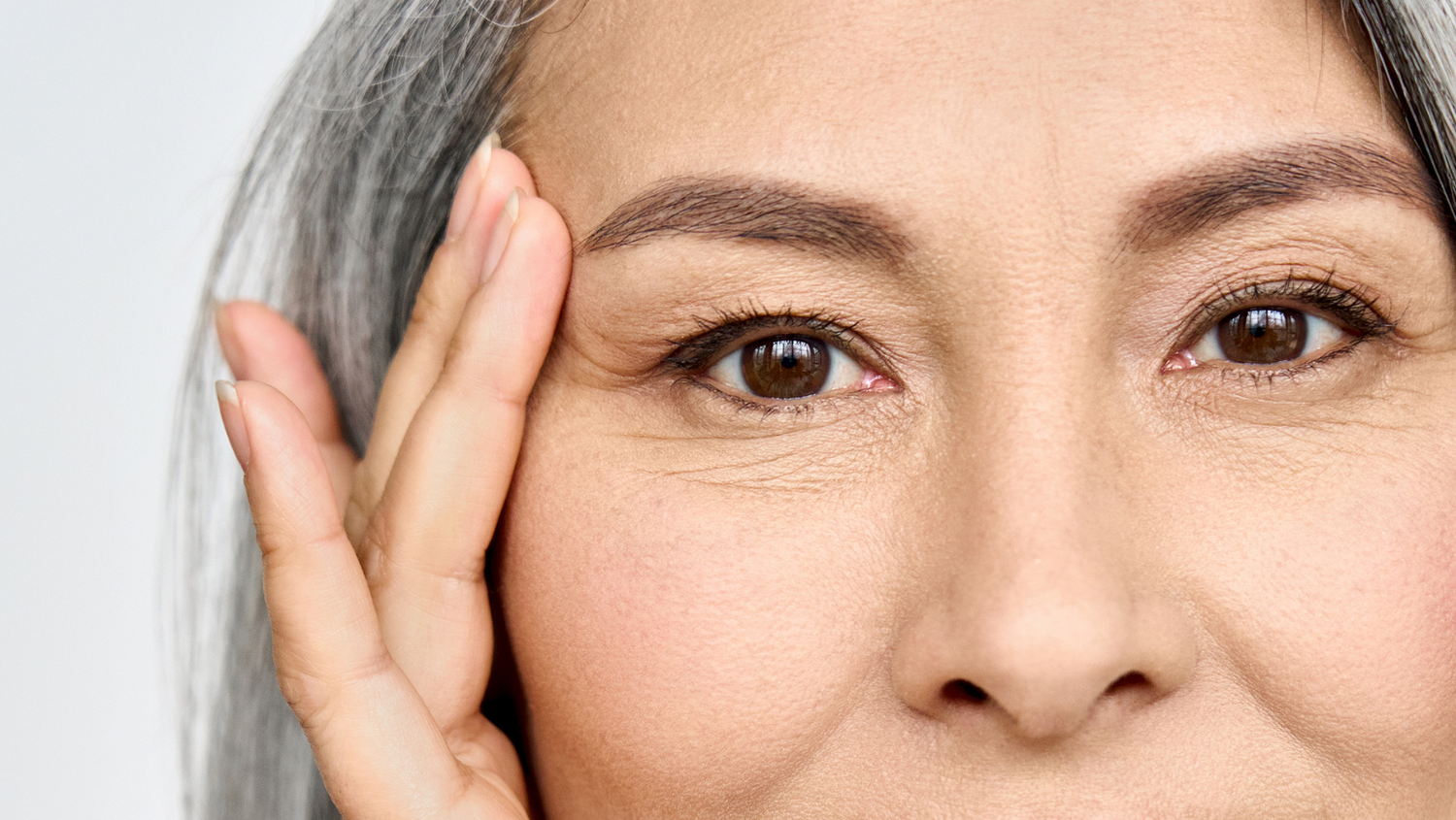 Eine ältere Frau mit grauen Haar streicht sich mit den Fingern über die Schläfe, ihre braunen Augen schauen den Betrachter an. 